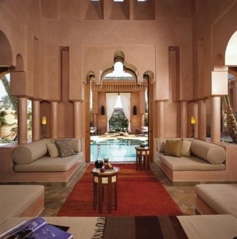 a warm-colored Moroccan patio wth carved sides and pillars, with bright rugs and blankets and comfy furniture