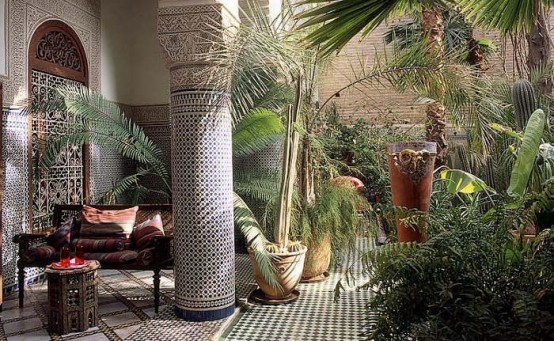 a chic Moroccan space with a tiled floor, a carved bench with bright upholstery and pillows and much potted greenery