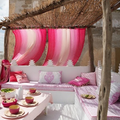a bright Moroccan patio done in pink and white, with colorful pillows and curtains and a low table