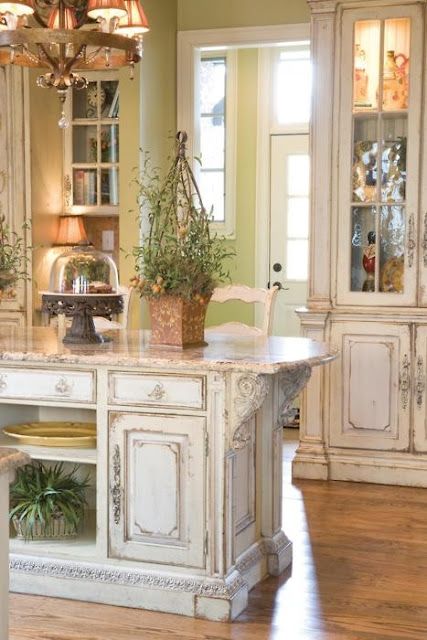 a refined shabby chic kitchen with neutral furniture, glass cabinets and a kitchen island, potted greenery and a vintage chandelier