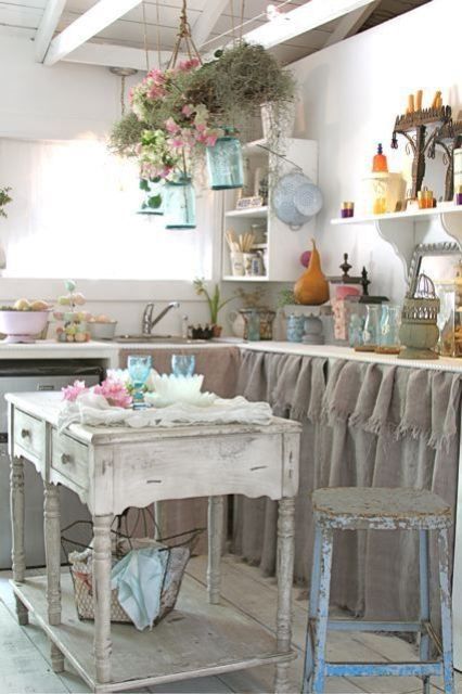 a rustic shabby chic kitchen with white walls, vintage furniture with burlap instead of doors, a shabby chic kitchen island and blooms hanging in jars over the island