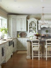 a refined shabby chic kitchen with neutral shabby cabinets, a crystal chandelier, a tan kitchen island with a black countertop
