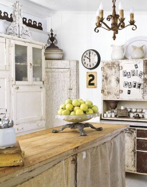 a shabby chic kitchen with neutral furniture, a kitchen island with burlap covering it, white cabinets, a vintage chandelier and some vintage decor