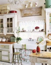 a bright shabby chic kitchen with olive green and white cabinets, a sink with a floral curtain, shabby chic wooden shelves, a crystal chandelier