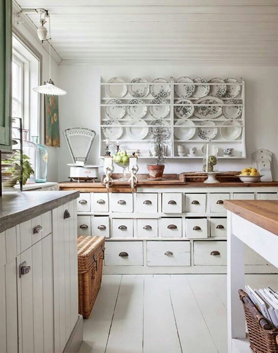 a white shabby chic kitchen with farmhouse cabinetry, a large display with floral plates, woven items and green touches