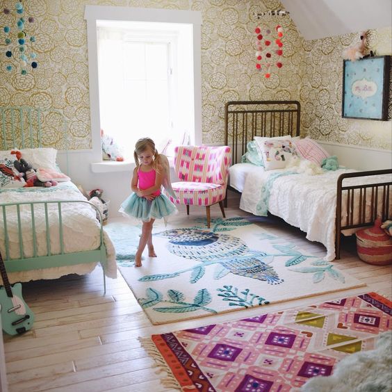 a bright and fun shared girls' bedroom with mismatching metal beds, white vintage bedding, colorful rugs, bright pompom mobiles and toys
