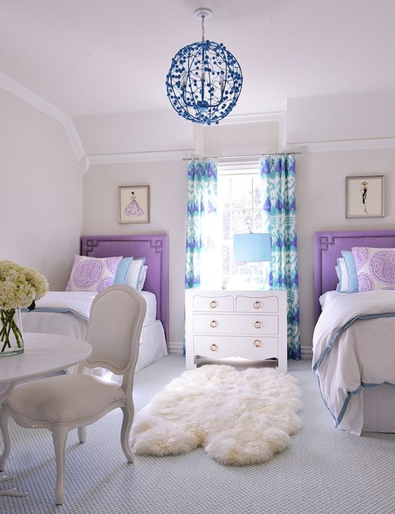 a refined shared girls' bedroom with purple upholstered beds, purple, white and blue bedding, a chic white dresser and a white table plus a chair, teal printed curtains