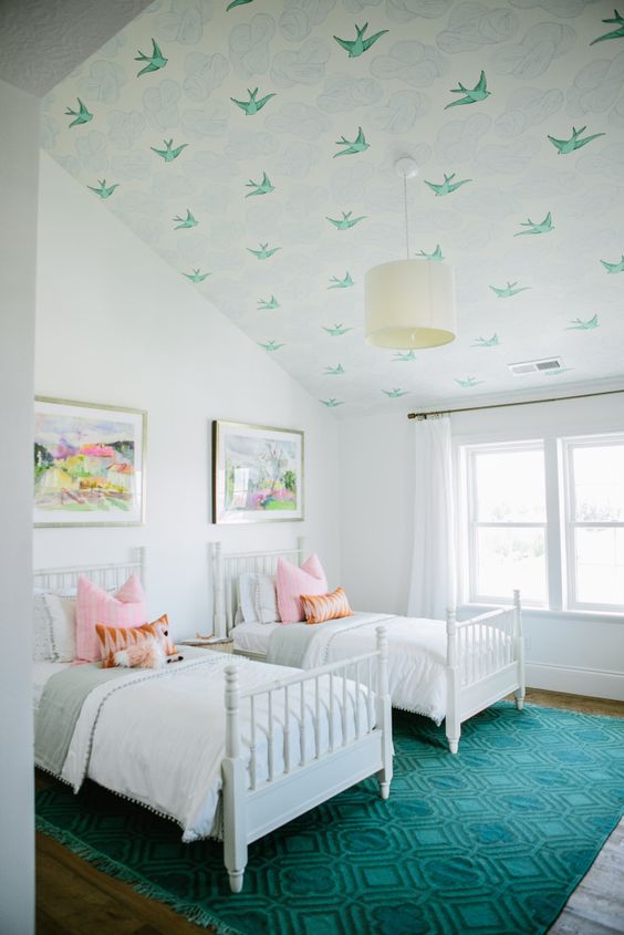 a dreamy white shared attic bedroom with a bird-printed ceiling, white vintage beds, neutral and pastel bedding, a teal rug and a bright artworks is cool