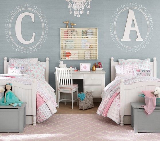 a shared girls' bedroom with a grey accent wall with monograms, white vintage beds and grey chests for storage, pink bedding, a white desk with a chair that doubles as a nightstand