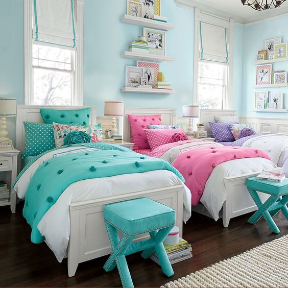 a pastel shared girls' bedroom with a light blue accent wall, three vintage neutral beds, purple, pink and turquoise bedding, turquoise stools, bookshelves on the walls