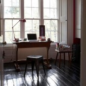 a vintage home office with a chic stained desk, a black stool, a table lamp, some signs, a stool with brushes and some drawers