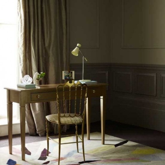 a moody home office with dark green walls with paneling, a small stained desk and a vintage chair, a table lamp and some books