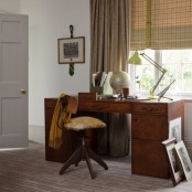 a vintage home office with a stained desk and a vintage chair, layered curtains, artwork, a green table lamp and some books and files