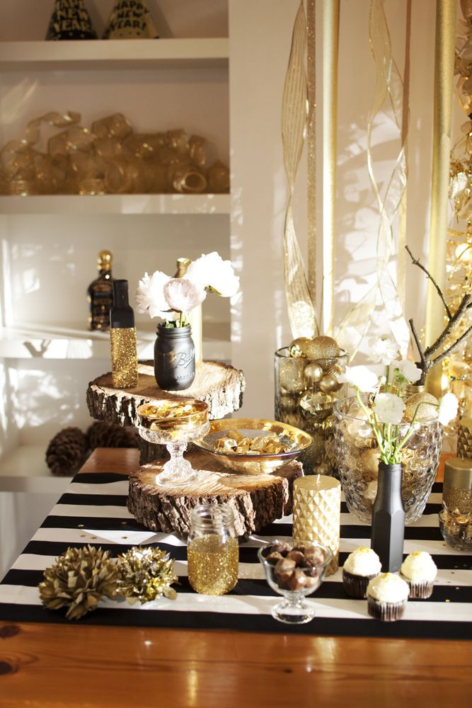 a glam NYE dessert table with a striped tablecloth, gold glitter bottles and candleholders, glasses with gold ornaments and various desserts