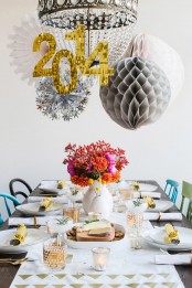 a bright NYE party tablescape with a printed table runner, white porcelain, a bright floral centerpiece, a chandelier decorated with paper pompoms and snowflakes and numbers of the year