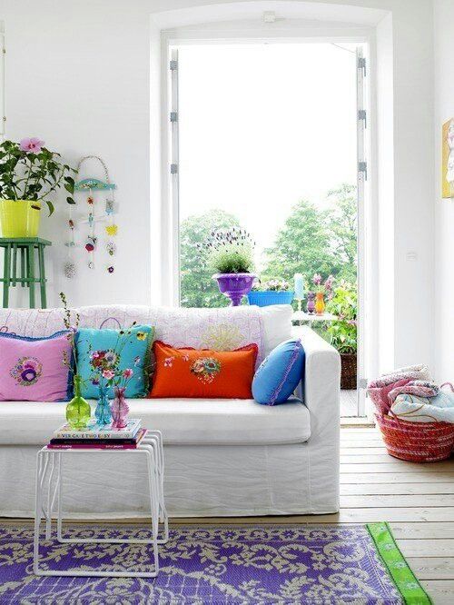 a bright and welcoming summer living room with bold floral pillows, with a colorful printed rug and some bold blooms in pots inside and outside, on the balcony