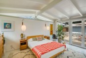 a bright mid-century modern bedroom with a warm colored bed and nightstands, rugs, artworks and windows for more light