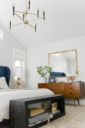 a chic and bright mid-century modern bedroom with white walls, rich stained furniture, a black storage bench and a gilded chandelier