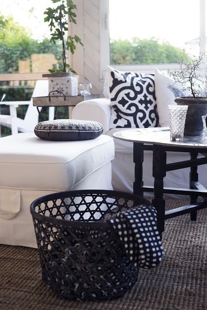 a porch with a white sectional, printed pillows, a black table and a black basket with blankets, greenery around