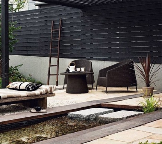 a stylish modern outdoor space with dark wicker chairs, a wooden daybed, printed pillows and a blanket