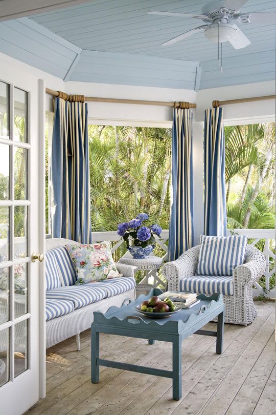 a beach sunroom with white wicker furniture and blue striped upholstery plus a vintage blue table and blue curtains