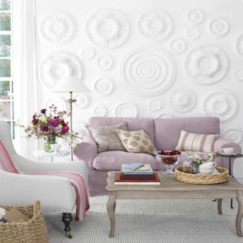 an airy living room with a decorative textural wall, a lilac sofa, a wooden table, some pink and lilac pillows and bold blooms