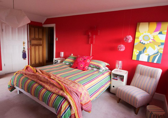 a super bright red bedroom with a bed covered with colorful striped bedding, a neutral chair and bold artwork and pillows is a lovely space to be in