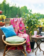 a bright and welcoming boho balcony with a rattan chair, a vintage side table, potted plants and blooms and colorful pillows and a blanket