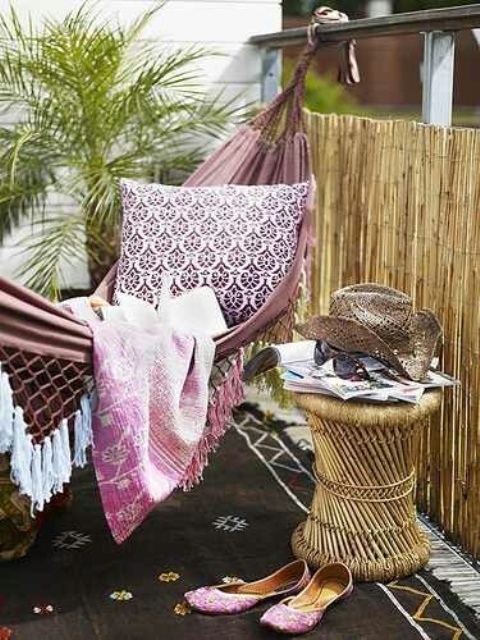 a boho balcony styled with a pink hammock, printed pillows and a blanket, a boho side table, some tropical plants and a black boho rug
