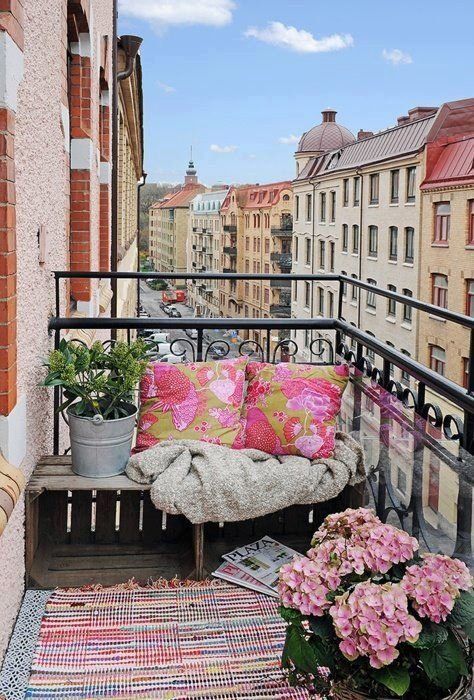 a colorful boho balcony with a bright printed pink rug and pillows on the wooden bench, some potted blooms and greenery is lovely and inviting