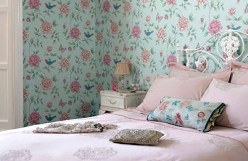 a pastel feminine bedroom with a blue and pink floral print wall, a bed with lilac and blue bedding, a vintage nightstand with a small table lamp