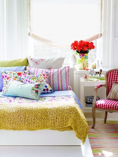 a neutral bedroom with white furniture and colorful bedding and pillows, a refined stained chair with checked red upholstery and bright blooms