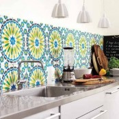 a neutral sleek kitchen with a grey countertop and a bold blue, green, yellow and black patterned tile backsplash that makes a bold accent to the space and pendant lamps