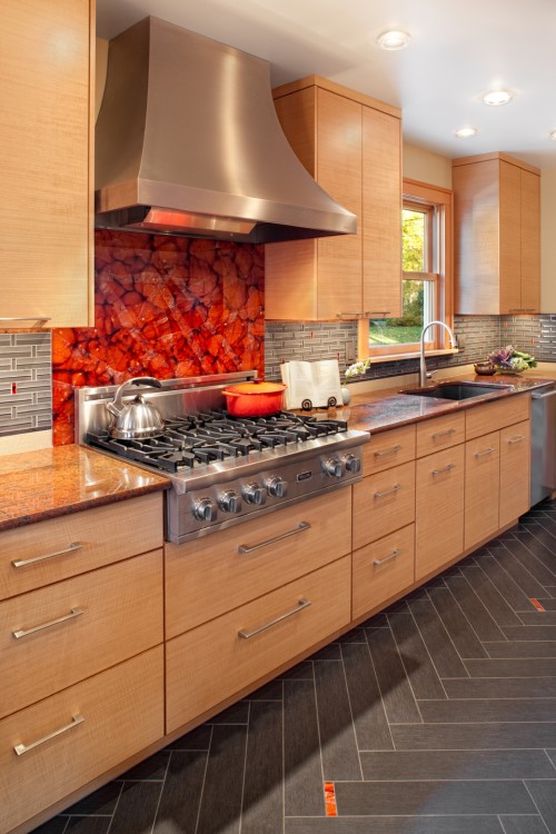 a light stained kitchen with modern handles and appliances plus a bold red glass backsplash over the cooker for a color accent in the space
