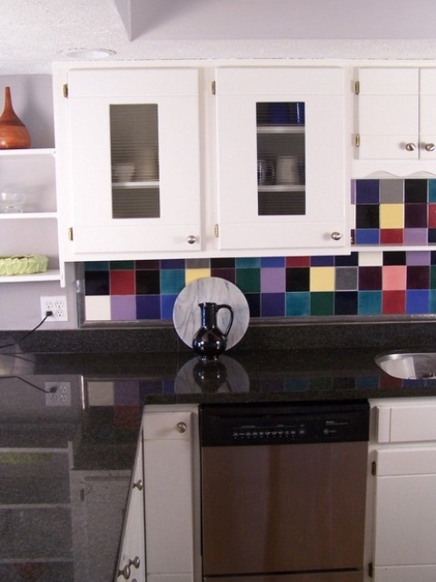 a white kitchen with dark grey countertops, a super colorful tile backsplash is a fun and bold idea wiht a cheerful mood