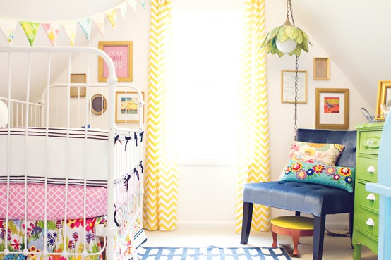 a colorful nursery with neutral walls, a white bed with colorful bedding, a green dresser, a navy chair, yellow printed curtains and a bold printed rug