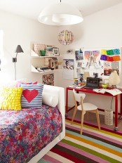 a neutral bedroom made bold mostly with textiles – bedding, a rug, pillows, with a red desk, a colorful gallery wlal and shelves with bright books