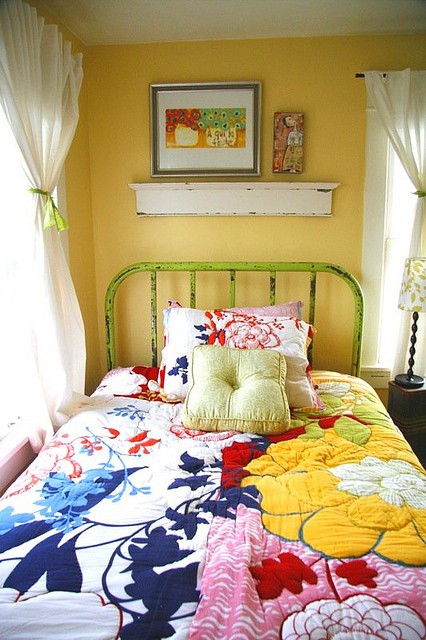 a colorful shabby chic bedroom with a yellow accent wall, a forged bed, colorful printed bedding, a small shelf with bright artwork is a cute nook to sleep in