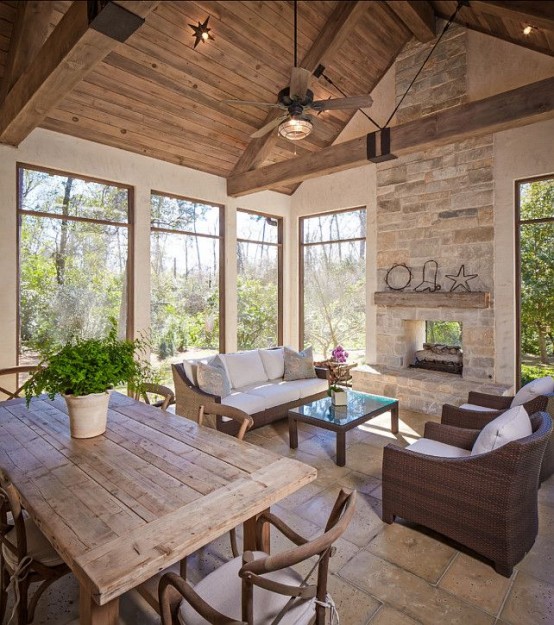 a neutral rustic screened porch with a fireplace, dark wicker furniture with neutral upholstery, a wooden coffee table and chairs, some greenery