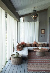 a lovely screened boho porch with a corner sofa with boho pillows, a boho rug, a side table and a copper coffee table plus white curtains for privacy and less light