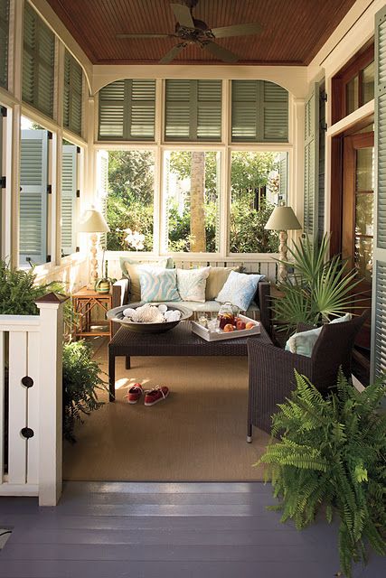 a screened porch with wicker furniture with striped upholstery, table lamps and greenery and green shutters on the windows