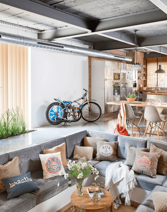 an industrial living room with a conversation pit, with a built-in sofa with grey upholstery, printed pillows and coffee tables and blooms