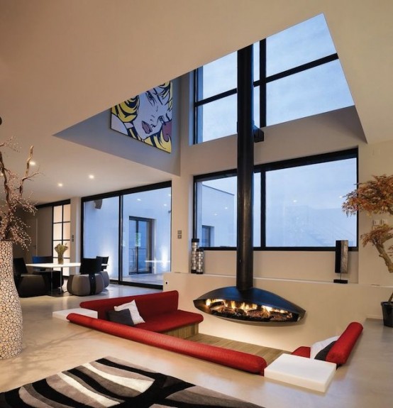 a modern living room with multiple windows, a conversation pit in front of one of them, a suspended black fireplace and red sofas in the pit