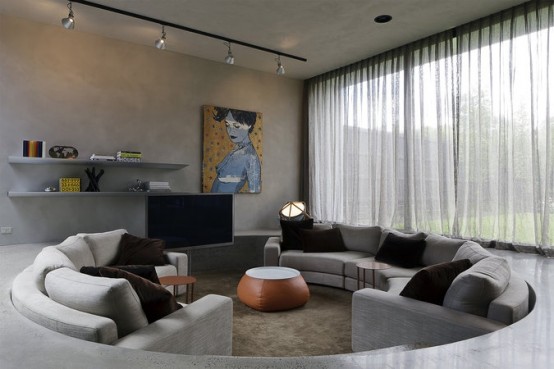 a contemporary living room of concrete with a round conversation pit, with grey sofas and black pillows plus a leather pouf in the center