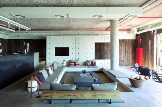 an industrial living room with a conversation pit with neutral sofas, lots of pillows and a large fire pit in the center