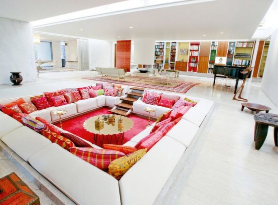 an all-white living room with a conversation pit, white sofas and bold and printed pillows, gold coffee tables and a bright red rug