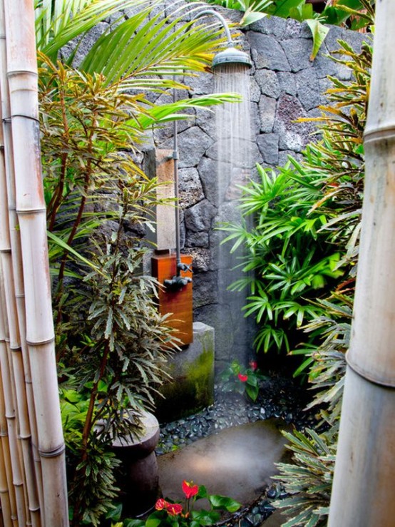 an outdoor tropical shower space with a stone wall and stones on the ground, tropical plants and a rain shower is a lovely idea