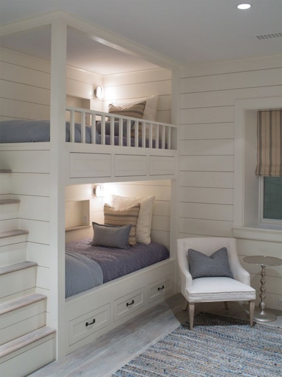 a white coastal bedroom with built-in bunk beds, with grey bedding, built-in lights and a chair with a grey pillow