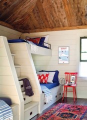 a nautical kids’ room with a wooden ceiling, built-in bunk beds with bright bedding, a colorful rug and bright artwork is a cool and lovely space