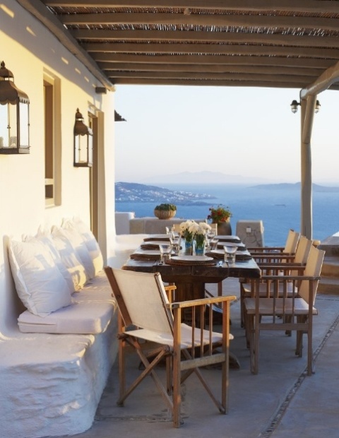 a Mediterranean summer terrace with a built-in bench, wooden furniture, candle lanterns and blooms and greenery plus a cool sea view